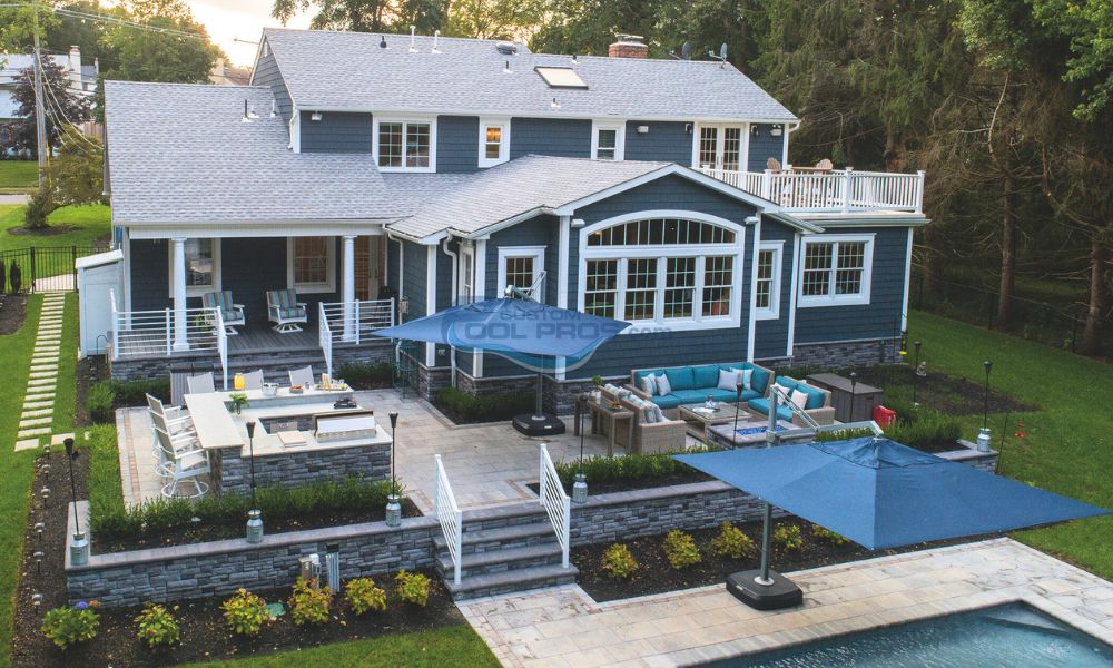 Modern blue house with a spacious outdoor patio, swimming pool, and lounge areas surrounded by landscaped gardens and umbrellas. Ideal for outdoor entertaining and relaxation.